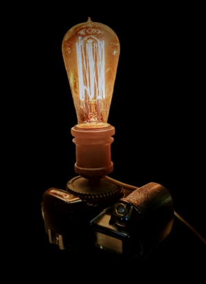 Lampa dekoracyjna z żarówką Edisona zamontowana w starym metalowym aparacie fotograficznym na drewnianym blacie.