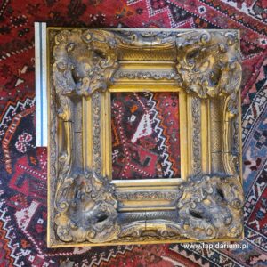 Masywna, zabytkowa rama dekoracyjna wykonana z drewna i gipsu na tle neutralnym.