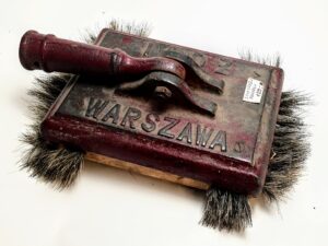 Zabytkowa żeliwna froterka ręczna z czerwonym korpusem i napisem WARSZAWA na białym tle.